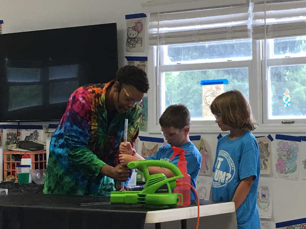 A member of Science Tellers wearing a bright tie dyed lab coat instructs two campers on an activity.