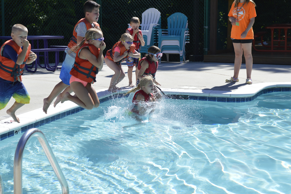 Children wearing life jackets jump into the Frog Pond Pool's crystal blue waters while a camp counselor wearing bright orange supervises.