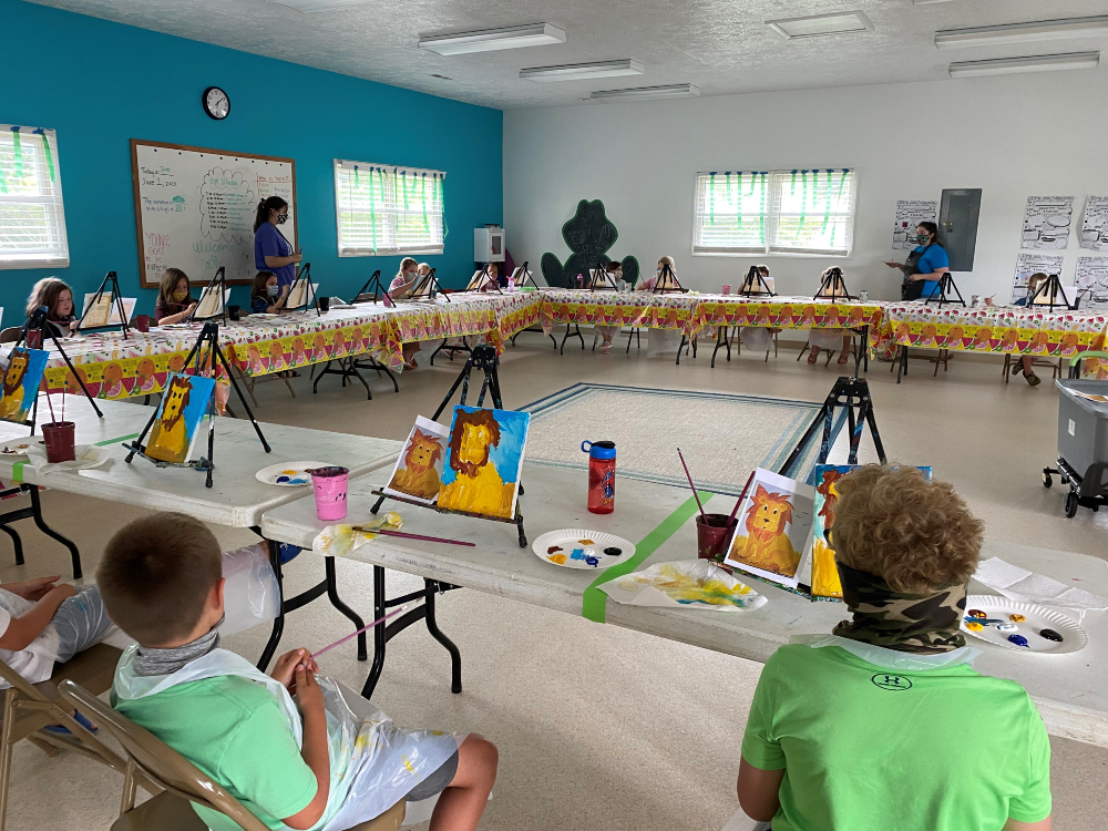 Children are spaced out at tables all around the Mid County Activity Center working on paintings of lions while camp counselors offer assistance.